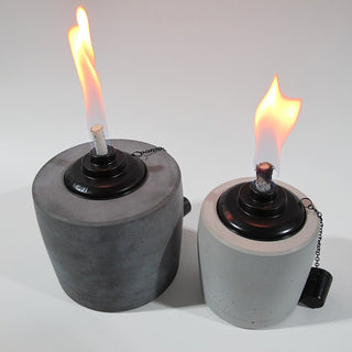 Links im Bild ist ein großes Betonfeuer in Anthrazit mit einer lang gezogenen Flame in schönem gelb/rot. Rechts daneben steht der kleine Bruder in der Farbe Grau, die Flame ist etwas breiter und scheint ebenso im schönen gelb/rot. Am unteren Rand der Gartenfackeln ist leicht das Branding der Marke Handworkz zu sehen, es ist in einem Guss mit dem Beton und somit sehr dezent und stilvoll.