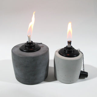 Links im Bild ein großes Betonfeuer in Anthrazit mit einer lang gezogenen Flame in schönem gelb/rot, rechts daneben steht der kleine Bruder in der Farbe Grau perfekt auf einer Höhe um den Größenunterschied der Betonfackeln schön zu sehen, die Flame ist schmal und scheint ebenso im schönen gelb/rot. Am unteren Rand der Gartenfackeln ist leicht das Branding der Marke Handworkz zu sehen, es ist in einem Guss mit dem Beton und somit sehr dezent und stilvoll.