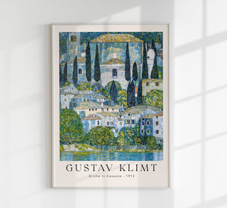 Exhibition-Poster zu Gustav Klimts „Kirche in Cassone“ mit dicht aneinanderliegenden Häusern, Kirche und Zypressen am Seeufer im quadratischen Format, in leuchtenden Grün- und Blautönen als Fine-Art-Print gerahmt an einer hellen Wand in einem modernen Wohnraum präsentiert
