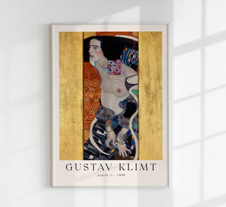 Ausstellungsplakat zu Gustav Klimts „Judith II (Salome)“ mit schlanker, dunkelhaariger Frau im vertikalen Format, tiefen Dunkeltönen, goldenen Ornamenten und typografischem Ausstellungstitel, als Fine-Art-Print gerahmt an einer dunklen Akzentwand in einem modernen Wohn- oder Galerieraum präsentiert
