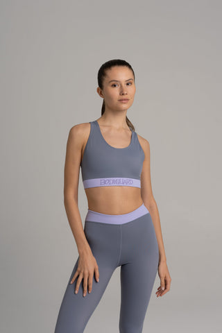 Grauer Sportsbra „Lagonisi“ mit lavendelfarbenem Unterbrustbund und rückseitiger Handytasche, getragen zu High-Waist-Leggings im Fitnessstudio.```

