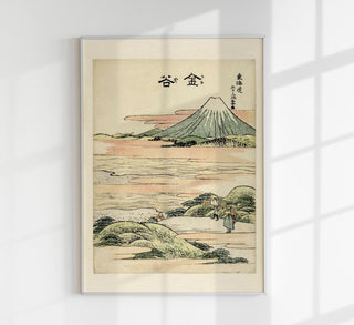 Japanischer Farbholzschnitt im Stil Hokusais mit Reisenden und Trägern, die einen breiten Fluss bei Kanaya überqueren, im Hintergrund Hügel und Berge unter hellem Himmel, als Fine-Art-Poster gerahmt an einer hellen Wand in einem modern eingerichteten Raum
