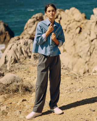 Graue Classic Hose für Damen aus zertifizierter Wolle mit Bundfalten, kombiniert mit Denimhemd, getragen vor Felsen am Meer.