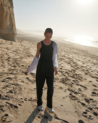 Schwarze Classic Hose für Damen aus zertifizierter Wolle, getragen mit schwarzem Top und offenem weißen Hemd beim Gehen am Strand.
