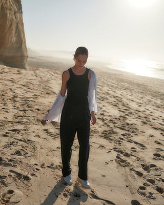 Schwarze Classic Hose für Damen aus zertifizierter Wolle, getragen mit schwarzem Top und offenem weißen Hemd beim Gehen am Strand.