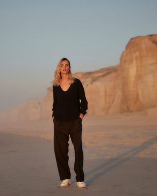 Schwarze Classic Hose für Damen aus zertifizierter Wolle, kombiniert mit schwarzem Pullover, getragen am Strand bei Sonnenuntergang.