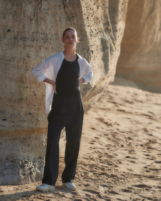 Schwarze Classic Hose für Damen aus zertifizierter Wolle, kombiniert mit schwarzem Top und weißem Hemd, getragen vor Felsen am Strand.