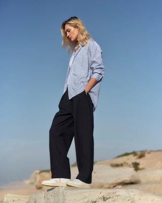 Schwarze Classic Hose für Damen aus zertifizierter Wolle, kombiniert mit blau-weiß gestreiftem Hemd, stehend auf einer Felsklippe am Strand.