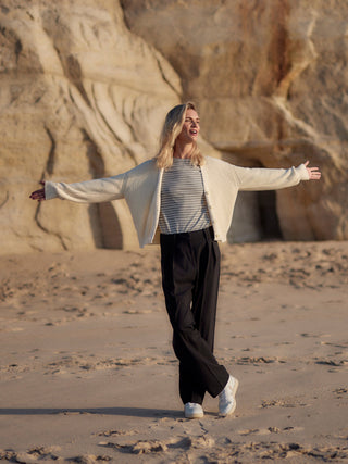Frau trägt eine schwarze Classic Hose aus zertifizierter Wolle am Strand, locker kombiniert mit gestreiftem Shirt und heller Strickjacke.