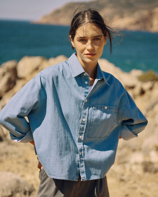Model trägt eine kurze hellblaue Denimbluse aus Bio-Baumwolle an einer Felsküste am Meer.