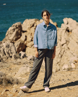 Kurze hellblaue Denimbluse aus Bio-Baumwolle, kombiniert mit weiter grauer Hose, getragen auf felsigem Gelände am Meer.