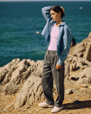 Kurze hellblaue Denimbluse aus Bio-Baumwolle, offen getragen über weißem Tanktop mit weiter grauer Hose, auf felsiger Küste am Meer.