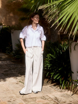 Frau trägt eine weiße, kurze Hemdbluse aus Bio-Baumwolle und weite, cremefarbene Hosen, stehend im sonnigen Garten mit Palmen und Steinboden.
