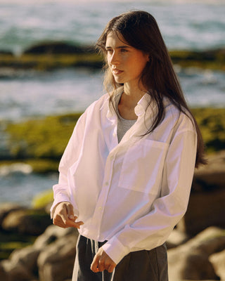 Kurze Damen Hemdbluse in Weiß aus Bio-Baumwolle, getragen am Meer im warmen Abendlicht, Seitenansicht.