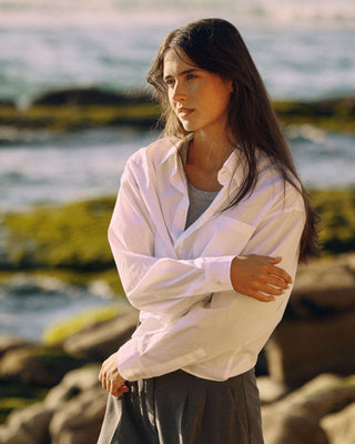 Kurze Damen Hemdbluse in Weiß aus Bio-Baumwolle, seitliche Ansicht am Felsenstrand im warmen Sonnenlicht.