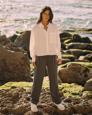 Kurze Damen Hemdbluse in Weiß aus Bio-Baumwolle, kombiniert mit grauer weiter Hose, stehend auf Felsen am Meer im warmen Sonnenlicht.