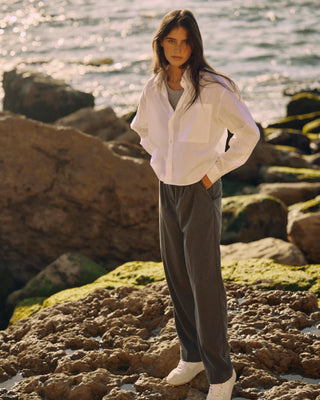 Kurze Damen Hemdbluse in Weiß aus Bio-Baumwolle, kombiniert mit grauer weiter Hose, stehend auf felsigem Untergrund am Meer im warmen Sonnenlicht.