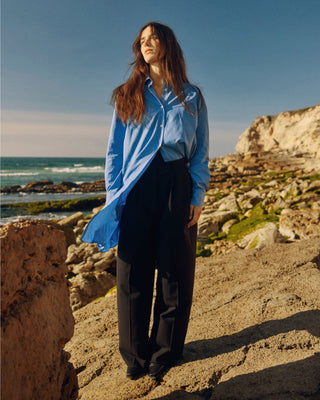 Schwarze Marlene Hose für Damen aus zertifizierter Wolle, kombiniert mit langer blauer Bluse, an einer felsigen Küste.