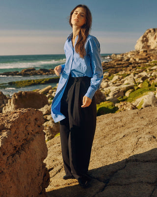 Schwarze Marlene Hose für Damen aus zertifizierter Wolle, kombiniert mit blauer Bluse, auf Felsen in einer Küstenlandschaft.