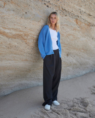 Schwarze Marlene Hose für Damen aus zertifizierter Wolle, kombiniert mit weißem Top und blauem Cardigan, getragen vor heller Felswand am Strand.