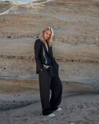 Schwarze Marlene Hose für Damen aus zertifizierter Wolle, kombiniert mit Oversize-Blazer, getragen vor heller Felswand.