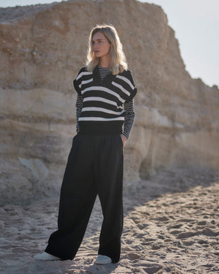 Schwarze Marlene Hose für Damen aus zertifizierter Wolle, kombiniert im gestreiften Layer-Look mit Langarmshirt und Strickweste, getragen am Strand vor Felskulisse.