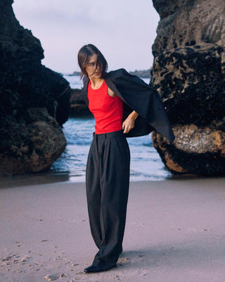 Schwarze Marlene Hose für Damen aus zertifizierter Wolle mit rotem Top, der schwarze Blazer lässig über die Schulter gelegt, am Strand in Bewegung.