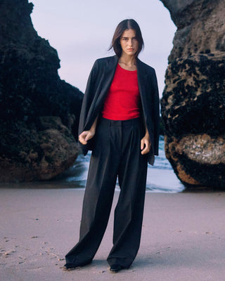 Schwarze Marlene Hose für Damen aus zertifizierter Wolle mit rotem Top und offenem schwarzem Blazer, frontal am Strand zwischen Felsen.