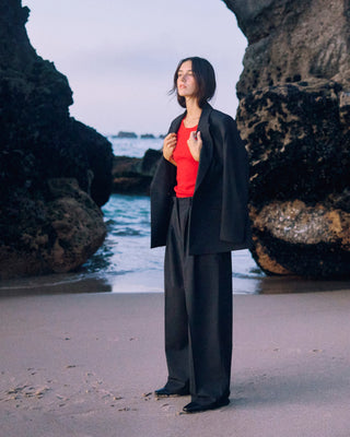 Schwarze Marlene Hose für Damen aus zertifizierter Wolle, kombiniert mit rotem Top und schwarzem Blazer, am Strand zwischen Felsen.