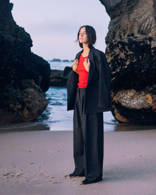 Schwarze Marlene Hose für Damen aus zertifizierter Wolle, kombiniert mit rotem Top und schwarzem Blazer, am Strand zwischen Felsen.