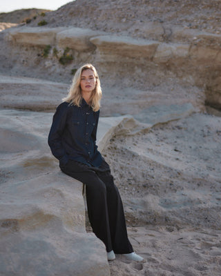 Schwarze Marlene Hose für Damen aus zertifizierter Wolle, kombiniert mit lockerem Denim-Hemd, sitzend in heller Felsenlandschaft am Strand.