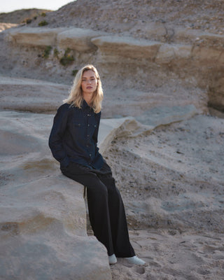 Schwarze Marlene Hose für Damen aus zertifizierter Wolle, kombiniert mit lockerem Denim-Hemd, sitzend in heller Felsenlandschaft am Strand.