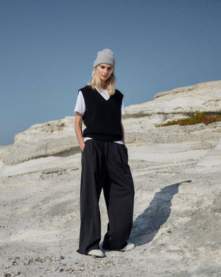 Schwarze Marlene Hose für Damen aus zertifizierter Wolle, kombiniert mit weißem T-Shirt, schwarzer Strickweste und grauer Mütze, getragen in heller Felslandschaft.