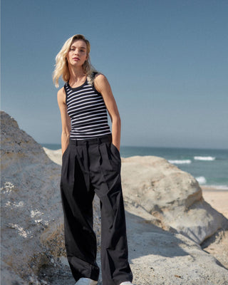 Schwarze Marlene Hose für Damen aus zertifizierter Wolle, kombiniert mit schwarz-weiß gestreiftem Top, getragen am sonnigen Steinstrand.