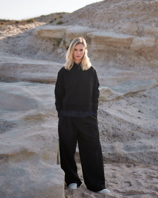Schwarze Marlene Hose für Damen aus zertifizierter Wolle, kombiniert mit schwarzem Sweatshirt, in heller Felsenlandschaft am Strand.