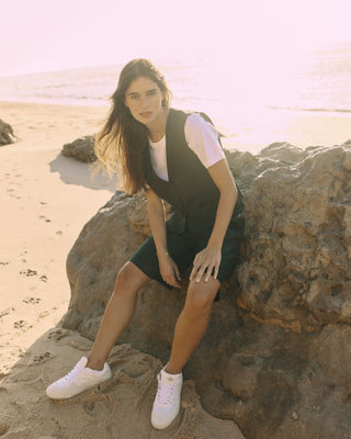 Model trägt eine schwarze Bermuda aus zertifizierter Wolle, sitzend auf einem Felsen am Strand.