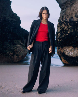 Model trägt schwarzen Damenblazer von WOTE aus nachhaltiger Tuchwolle über rotem Top und weiter Hose, stehend am Strand zwischen Felsen.