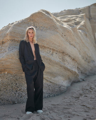 Model trägt schwarzen Damenblazer von WOTE aus nachhaltiger Tuchwolle mit weiter Hose, fotografiert am Strand vor heller Felsformation.