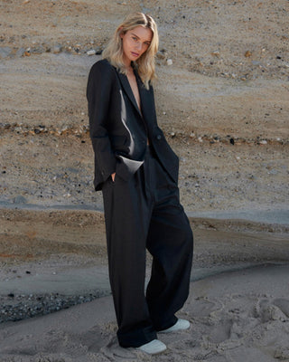 Model trägt schwarzen Damenblazer von WOTE aus nachhaltiger Tuchwolle mit weiter Hose, fotografiert vor geschichteter Sandsteinwand am Strand.