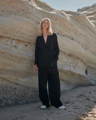 Model trägt schwarzen Damenblazer von WOTE aus nachhaltiger Tuchwolle mit weiter Hose, fotografiert vor heller Sandsteinformation am Strand.