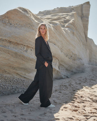 Model trägt schwarzen Damenblazer von WOTE aus nachhaltiger Tuchwolle und weiter Hose, gehend am hellen Sandstrand vor geschichteten Felsformationen.