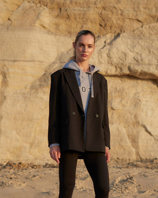 Model kombiniert schwarzen Damenblazer von WOTE aus nachhaltiger Tuchwolle mit grauem Hoodie, lässiges Layering vor heller Klippenwand im warmen Abendlicht.