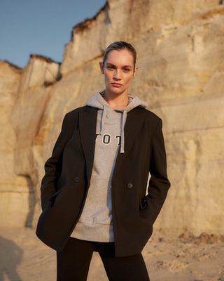 Model kombiniert schwarzen Damenblazer von WOTE aus nachhaltiger Tuchwolle mit grauem Hoodie, lässiges Layering vor sandfarbener Felswand im warmen Abendlicht.