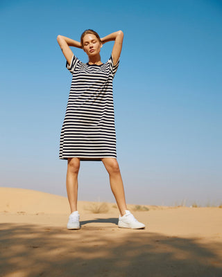 Oversized Damen T-Shirt-Kleid in Schwarz-Offwhite gestreift aus Bio-Baumwolle, getragen im Sand vor blauem Himmel.