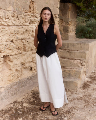 Schwarze WOTE Damenweste aus Tuchwolle, kombiniert mit langem weißem Rock, als Ganzkörperlook vor einer Natursteinwand fotografiert.