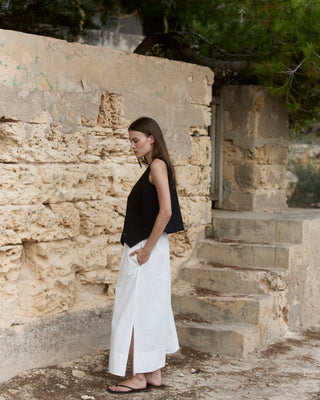 Seitliche Ansicht einer schwarzen WOTE Damenweste aus hochwertiger Tuchwolle, kombiniert mit fließendem weißem Maxirock, getragen vor einer hellen Natursteinwand.