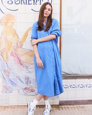 Model trägt ein einfarbig blaues Hemdblusenkleid für Damen in Midi-Länge aus Organic Cotton mit durchgehender Knopfleiste und lockerem Schnitt.