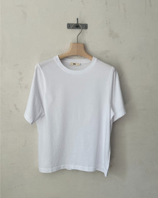 Weißes Relaxed-Fit Rundhals T-Shirt aus Bio-Baumwolle für Damen, aufgehängt an einem Holzkleiderbügel vor heller Wand.