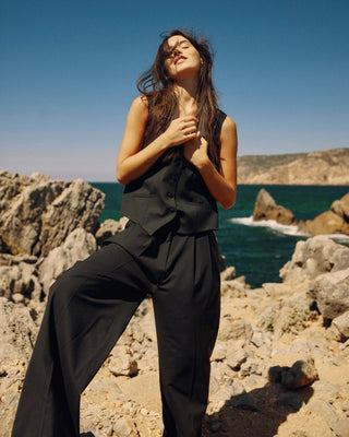 Model trägt eine schwarze Weste aus nachhaltiger Tuchwolle mit weiter schwarzer Hose auf felsiger Küste mit Meerblick.